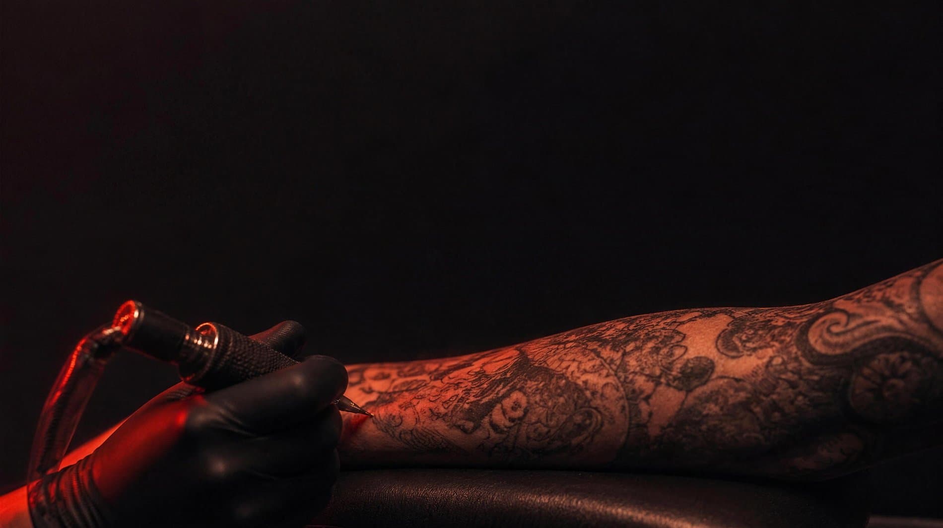 A gloved hand working a rotary tattoo machine across a heavily tattooed forearm under a single warm vermillion light
