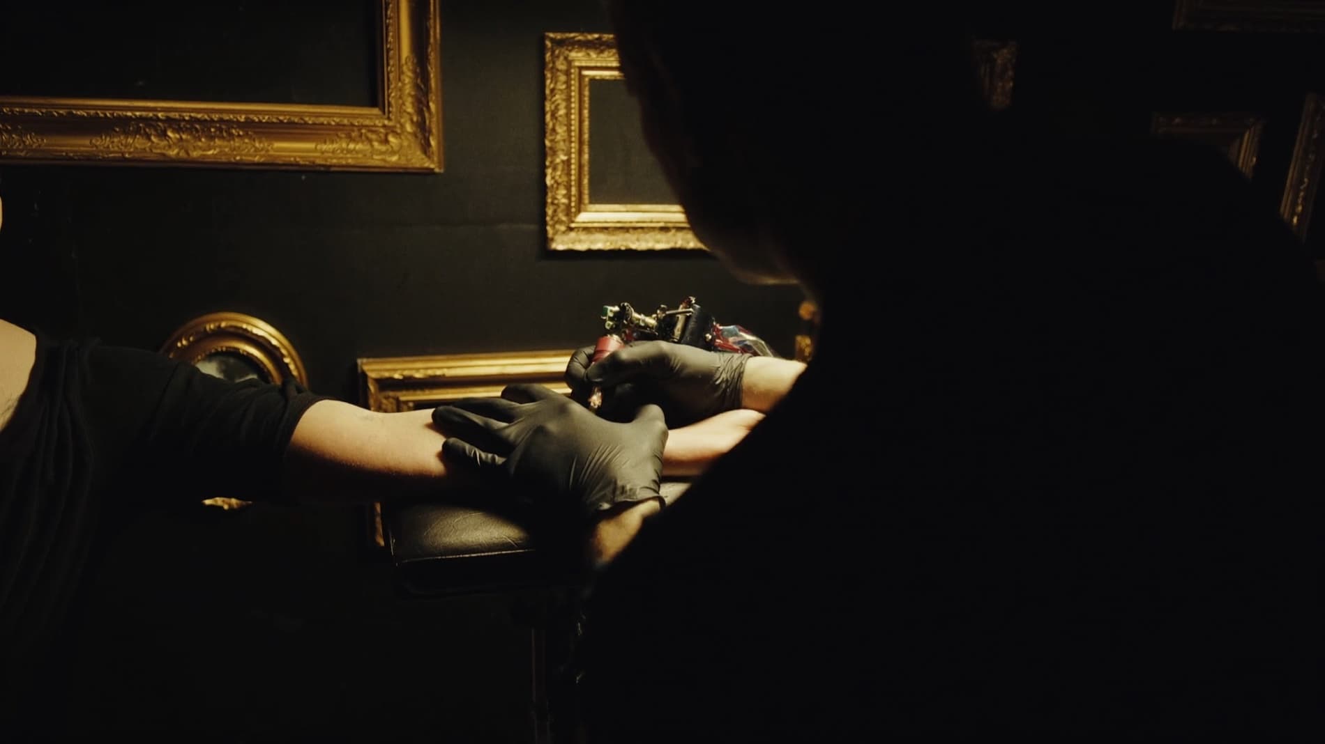 Tattoo artist at work in a dark gallery-style studio with gold-framed art